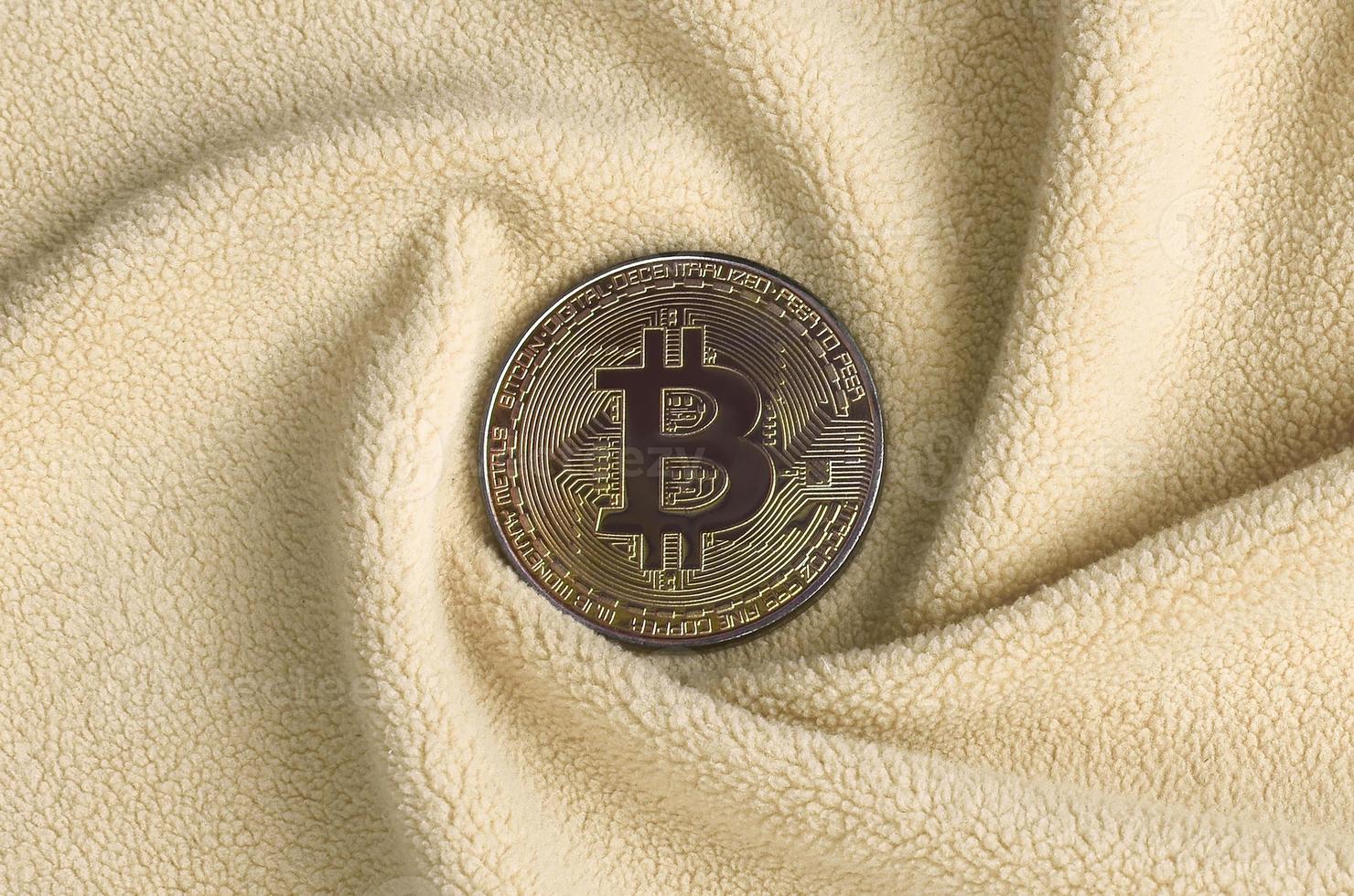 The golden bitcoin lies on a blanket made of soft and fluffy light orange fleece fabric with a large number of relief folds. The shape of the folds resembles a fan from a video card cooler photo