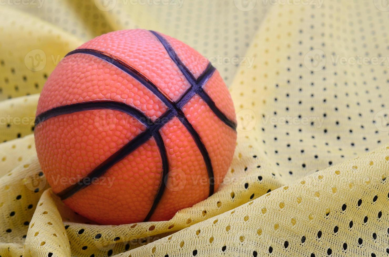 Small orange rubber basketball lies on a yellow sport jersey clothing fabric texture and background with many folds photo