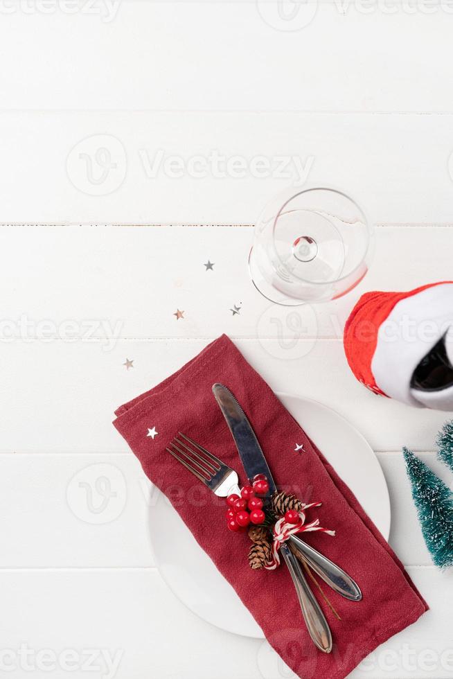 Christmas table setting with white dishware, silverware and red and green decorations on white wooden background. Top view. photo