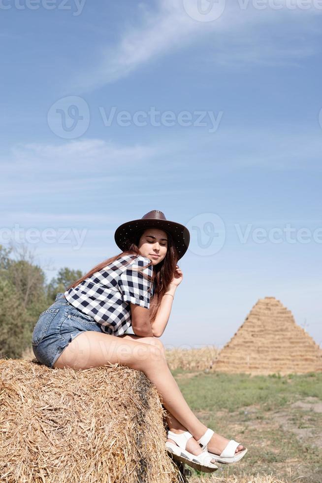 beautiful woman in plaid shirt and cowboy hat resting on haystack against sky background photo