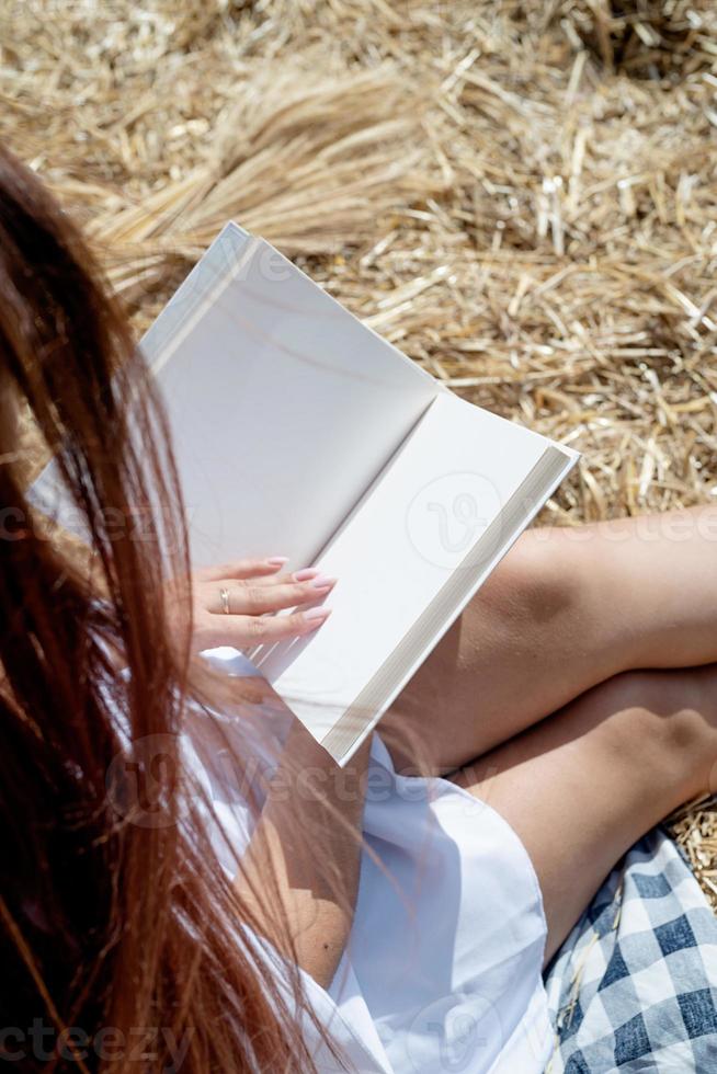 Young woman in white dress sitting on haystack in harvested field, reading blank book. Book mockup photo