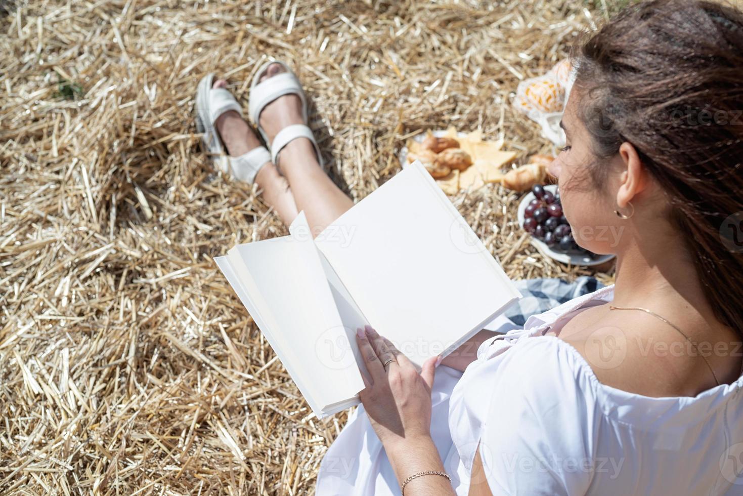 Young woman in white dress sitting on haystack in harvested field, reading blank book. Book mockup photo