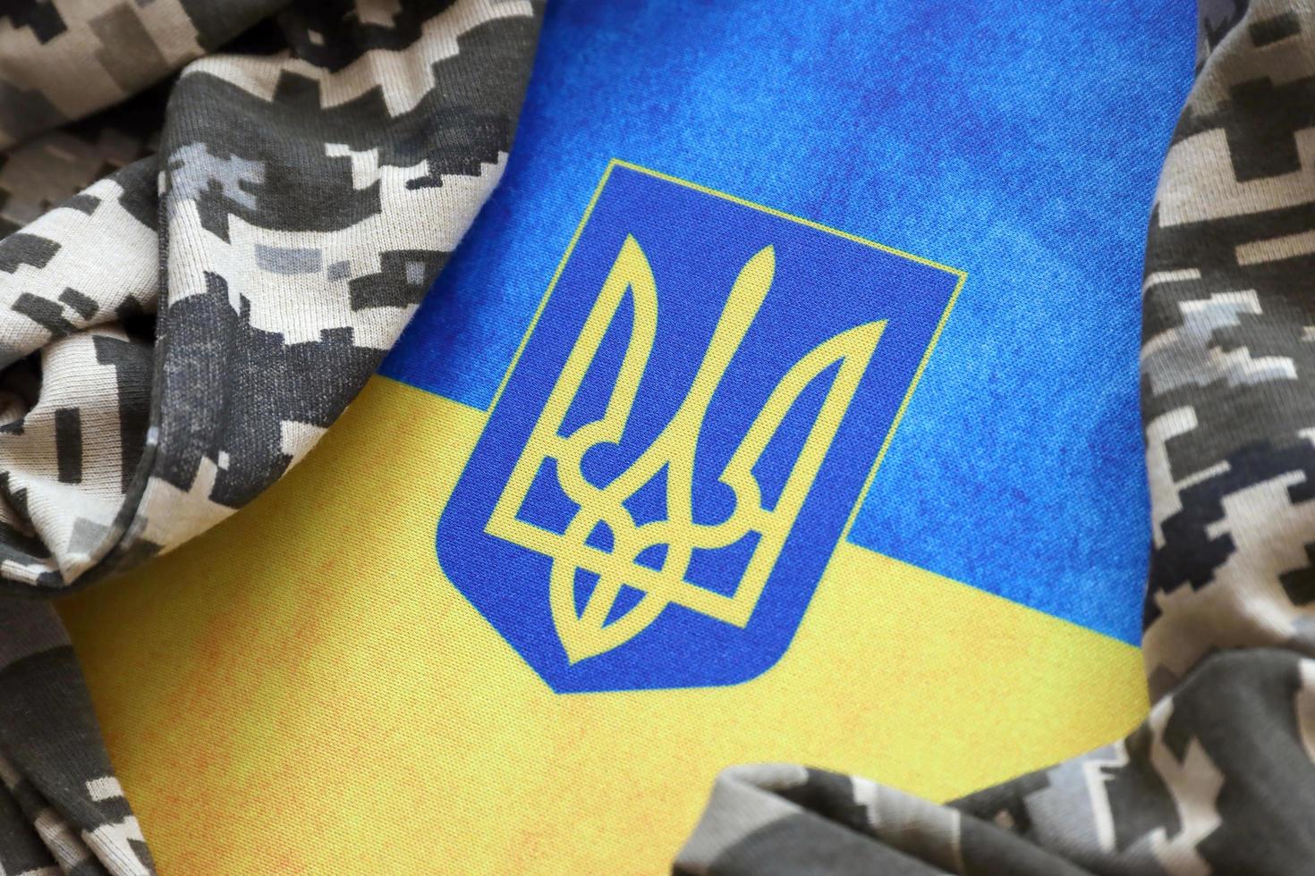 SUMY, UKRAINE - MARCH 20, 2022 Ukrainian flag and coat of arms with fabric with texture of pixeled camouflage. Cloth with camo pattern in grey, brown and green pixel shapes with Ukrainian trident sign photo