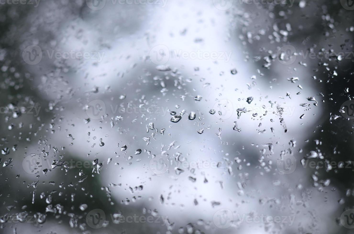 A photo of rain drops on the window glass with a blurred view of the blossoming green trees. Abstract image showing cloudy and rainy weather conditions