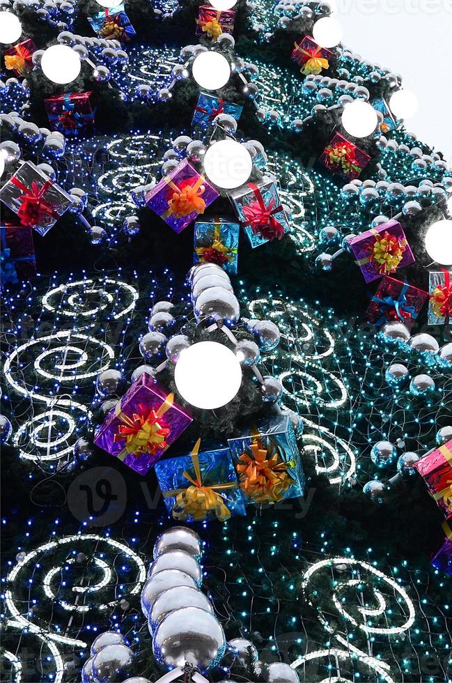 A fragment of a huge Christmas tree with many ornaments, gift boxes and luminous lamps. Photo of a decorated Christmas tree close-up