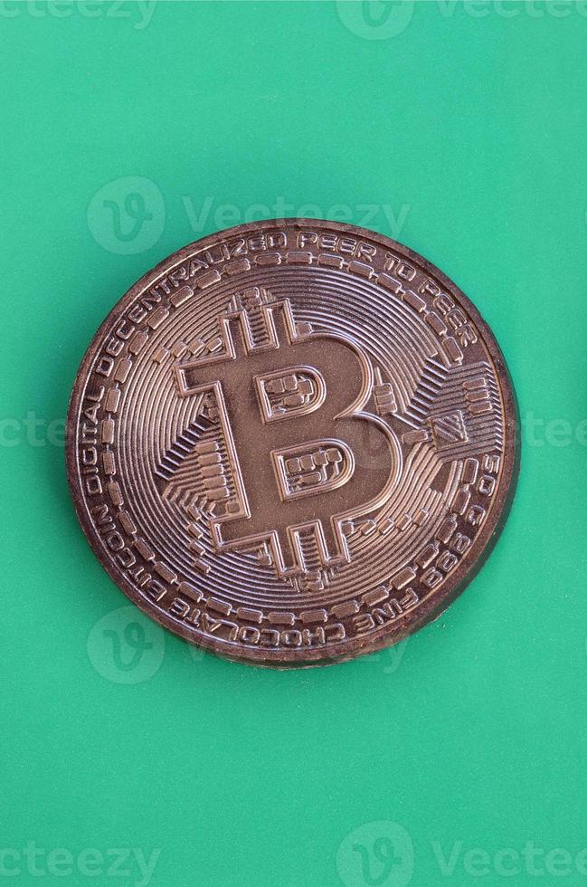 Chocolate product in the form of physical bitcoin lies on a green plastic background. Model of the crypto currency in the edible form photo