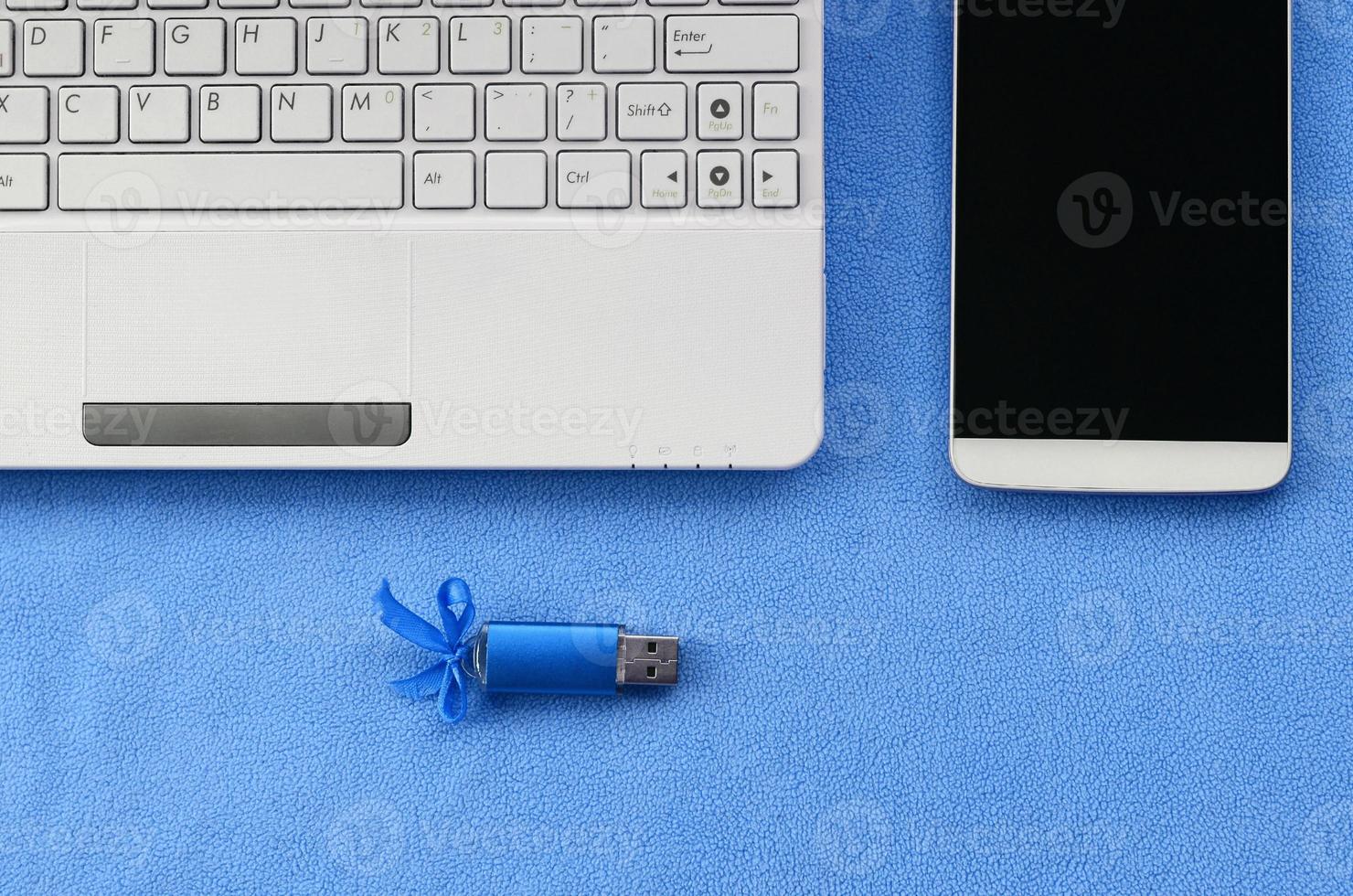 Brilliant blue usb flash memory card with a blue bow lies on a blanket of soft and furry light blue fleece fabric beside to a white laptop and smartphone. Classic female gift design for a memory card photo