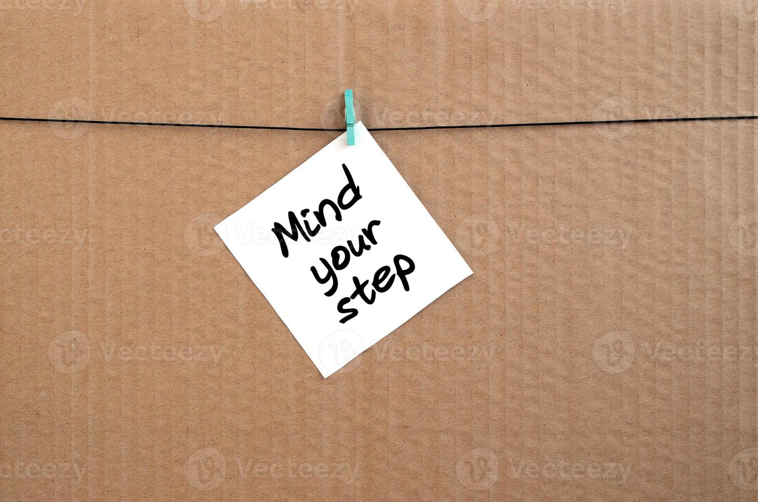 Mind your step. Note is written on a white sticker that hangs with a clothespin on a rope on a background of brown cardboard photo