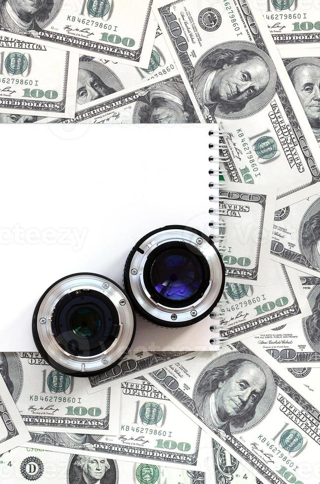 Two photographic lenses and white notebook lie on the background of a lot of dollar bills. Space for text photo