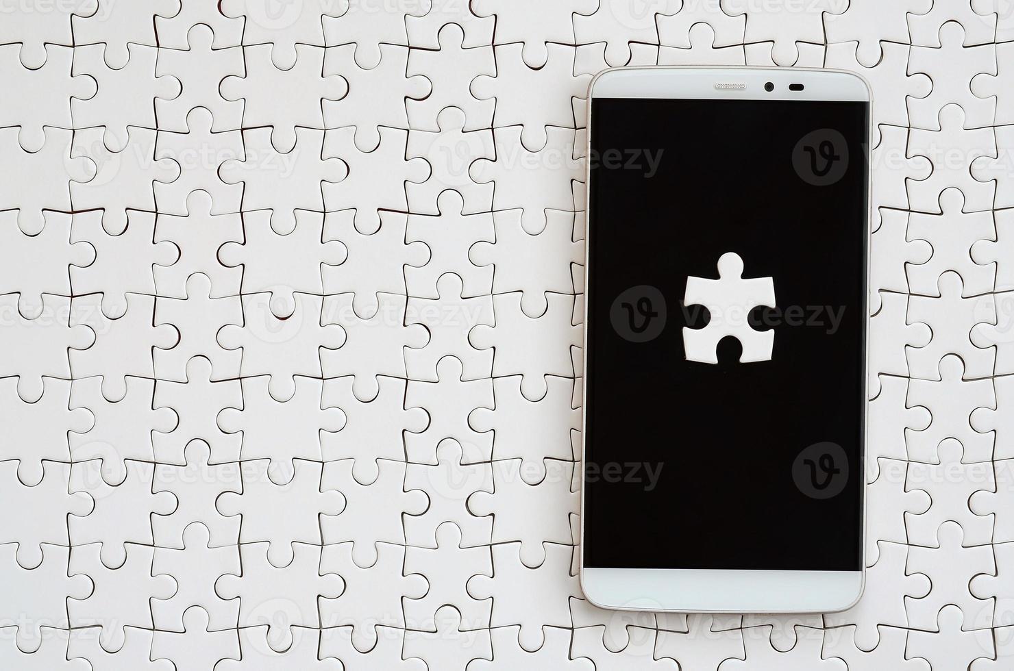 A modern big smartphone with several puzzle elements on the touch screen lies on a white jigsaw puzzle in an assembled state photo
