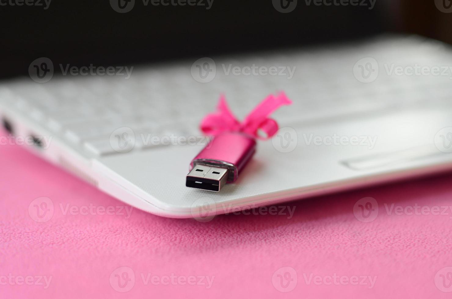 Brilliant pink usb flash memory card with a pink bow lies on a blanket of soft and furry light pink fleece fabric beside to a white laptop. Classic female gift design for a memory card photo