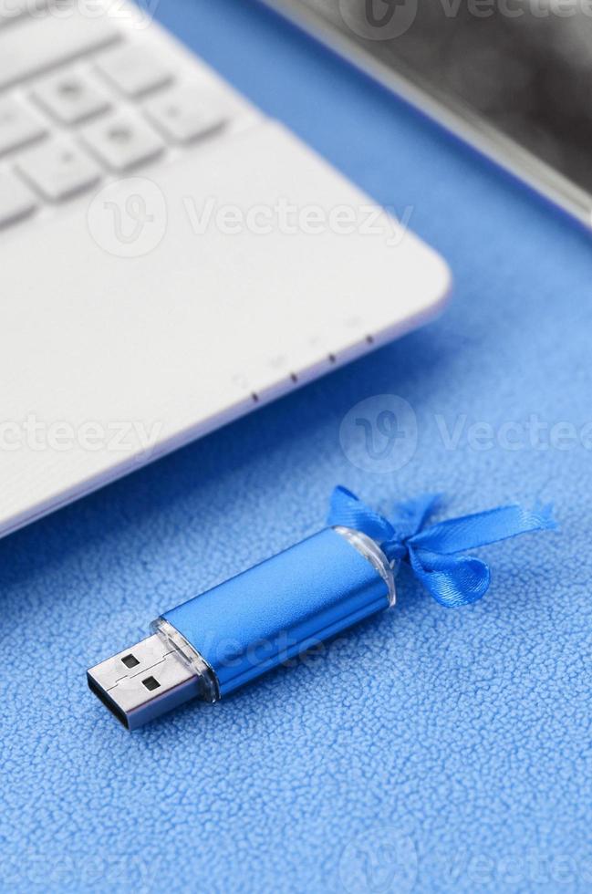 Brilliant blue usb flash memory card with a blue bow lies on a blanket of soft and furry light blue fleece fabric beside to a white laptop and smartphone. Classic female gift design for a memory card photo