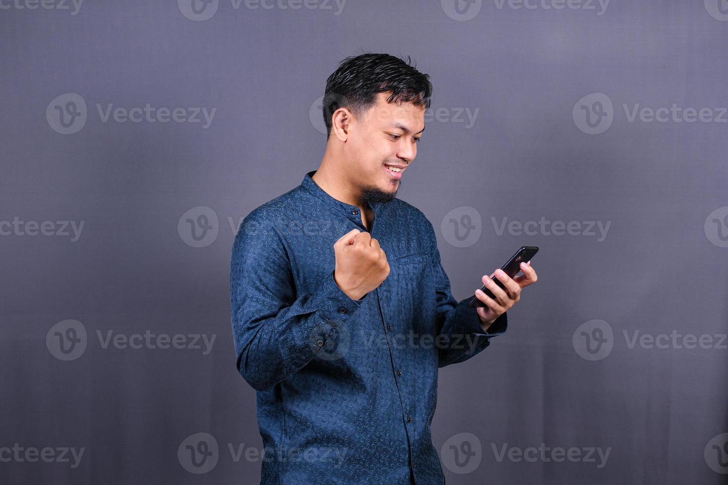 Happy young man in blue shirt posing isolated on gray background studio portrait. People lifestyle concept. Mock up copy space. Hold mobile phone, doing winner gesture photo