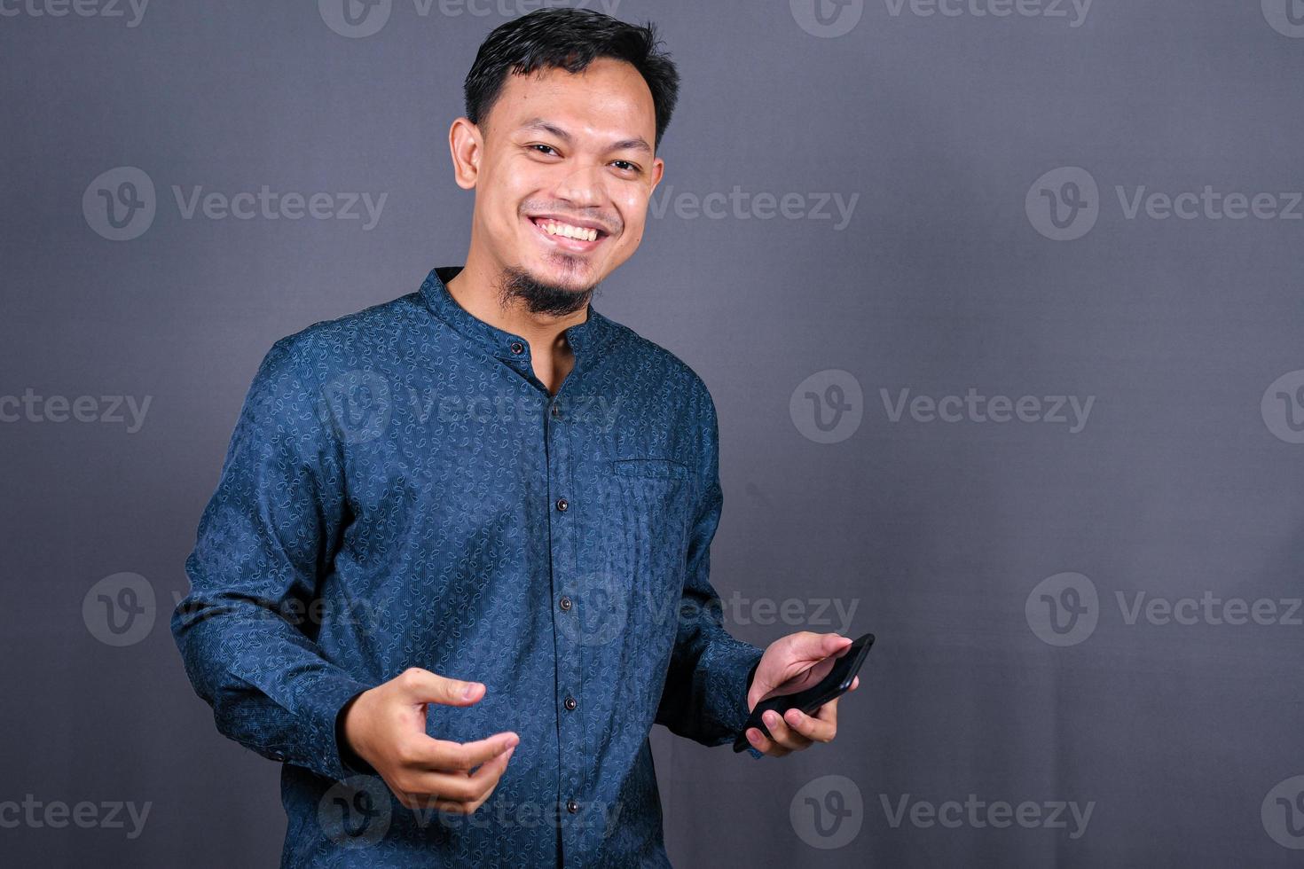 Happy young man in blue shirt posing isolated on gray background studio portrait. People lifestyle concept. Mock up copy space. Hold mobile phone, doing winner gesture photo