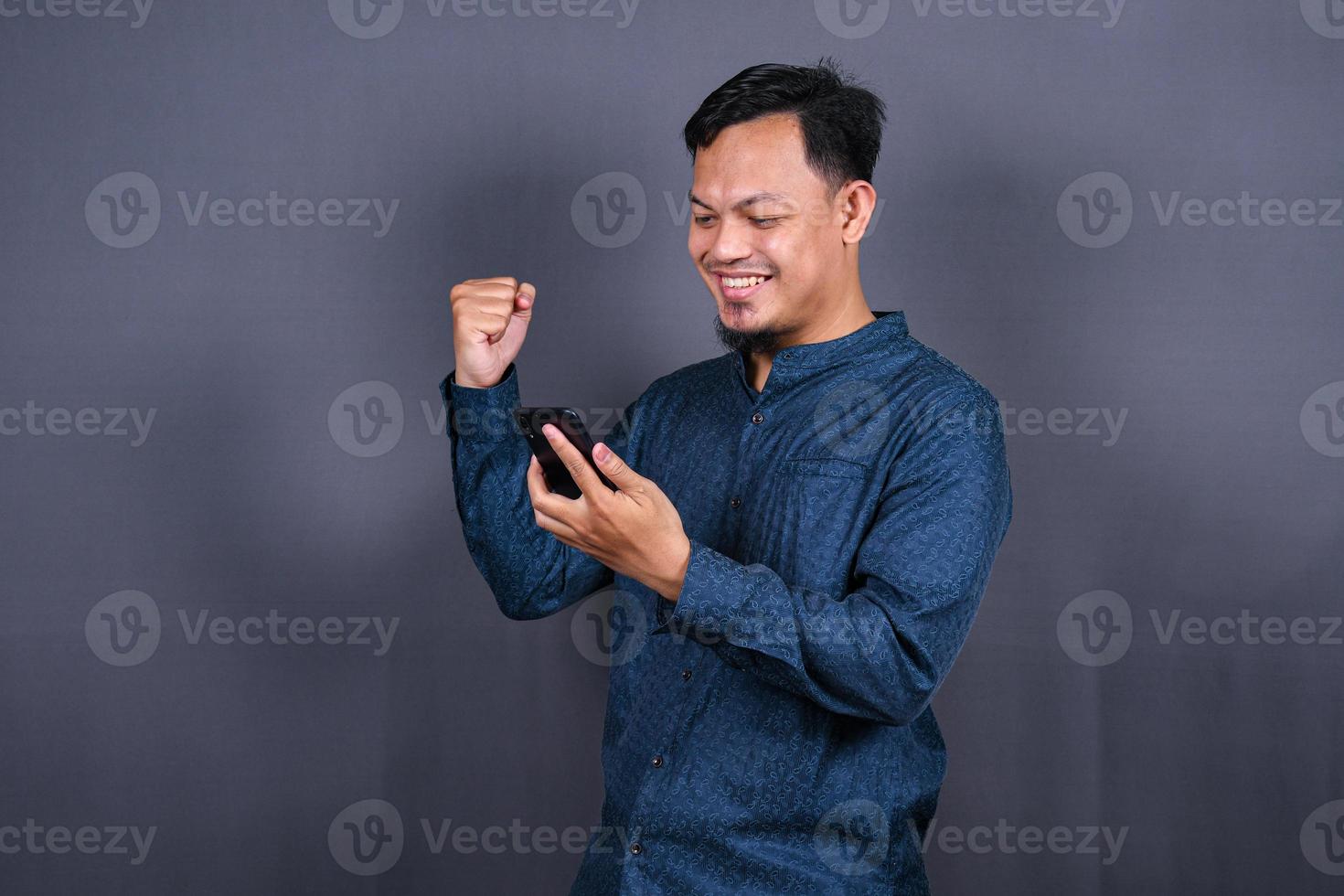 Happy young man in blue shirt posing isolated on gray background studio portrait. People lifestyle concept. Mock up copy space. Hold mobile phone, doing winner gesture photo