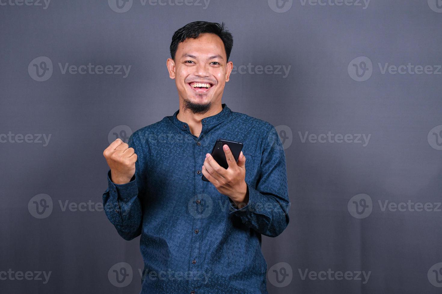 Happy young man in blue shirt posing isolated on gray background studio portrait. People lifestyle concept. Mock up copy space. Hold mobile phone, doing winner gesture photo