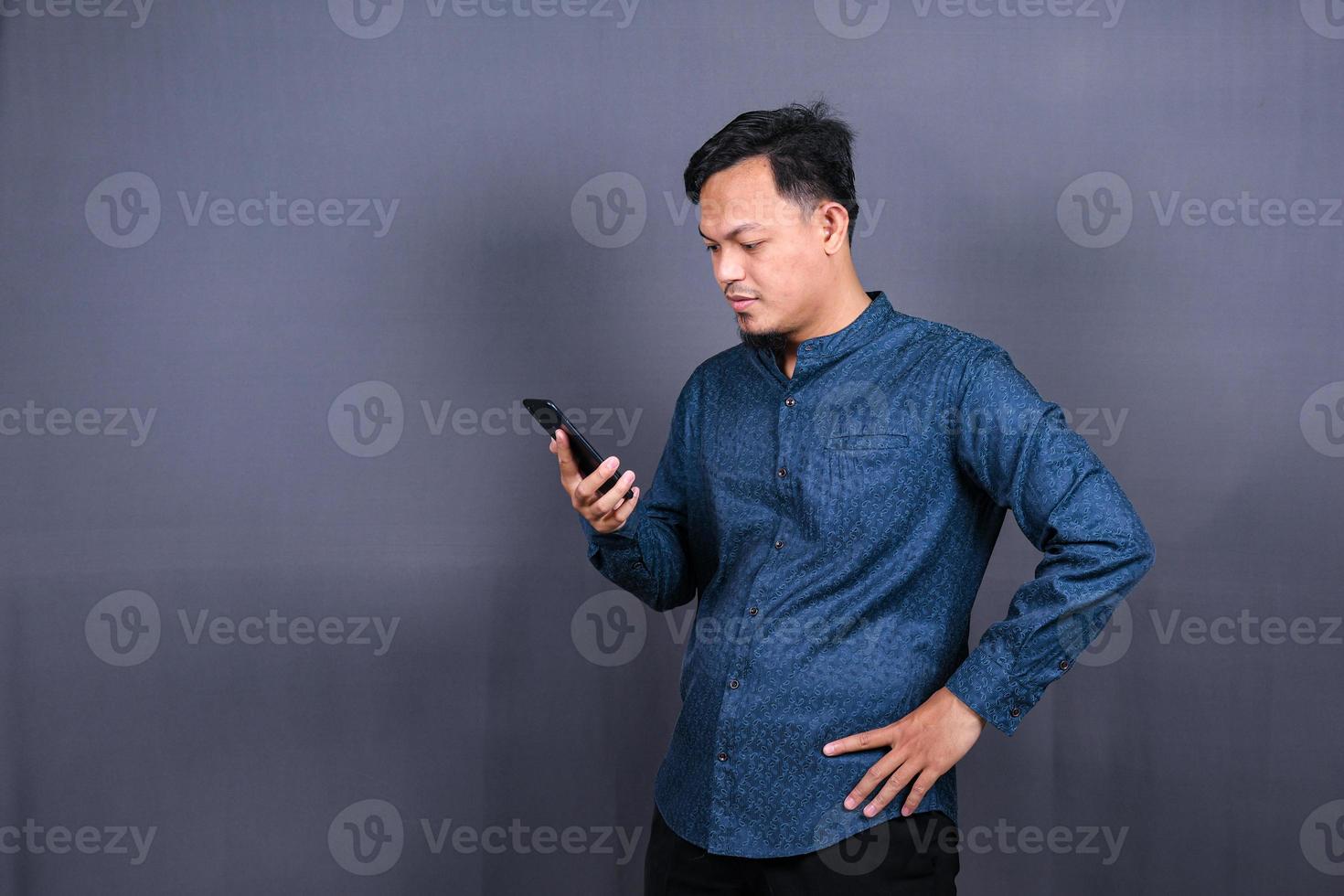 Portrait of unhappy Asian man in blue shirt holding mobile phone with sad expression on face. Isolated on gray background photo