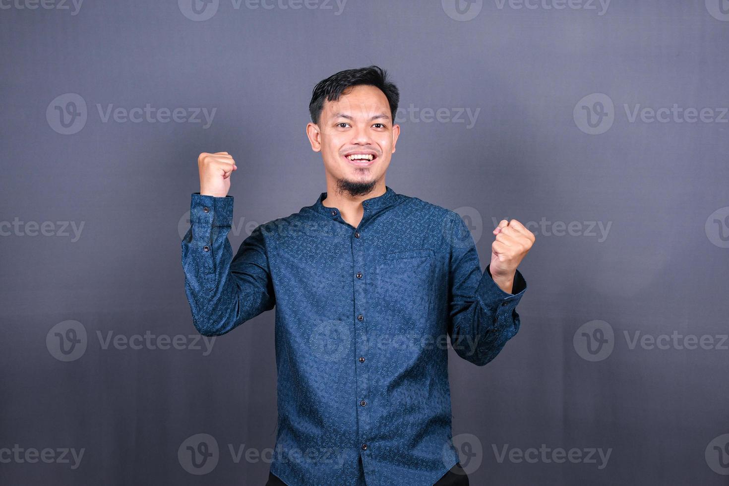 Young handsome man with blue shirt over gray background very happy and excited doing winner gesture with arms raised, smiling and screaming for success. Celebration photo