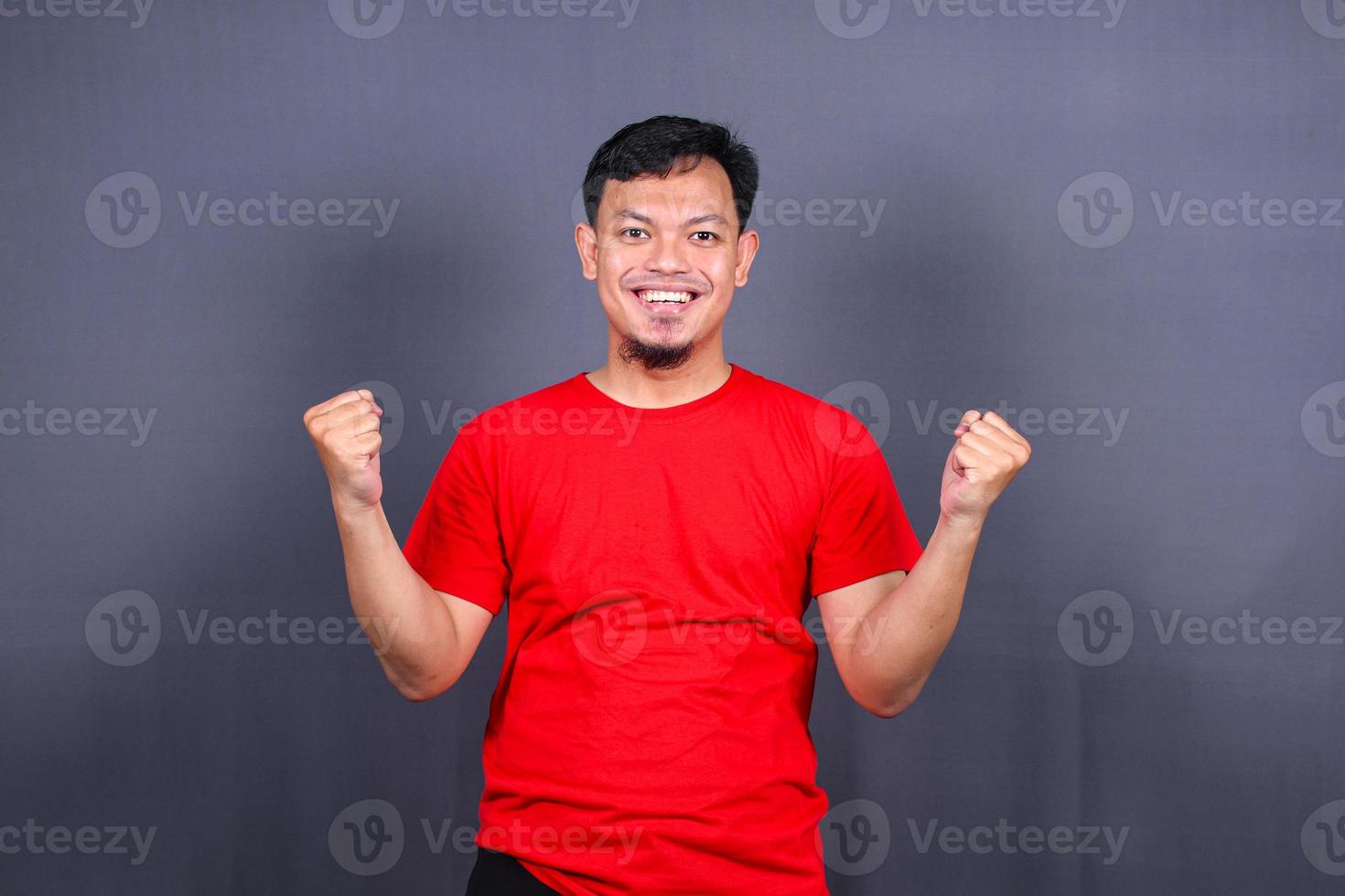 Portrait of an excited asian man standing with raised hands and looking at camera isolated on gray background celebrating succes, victory. photo