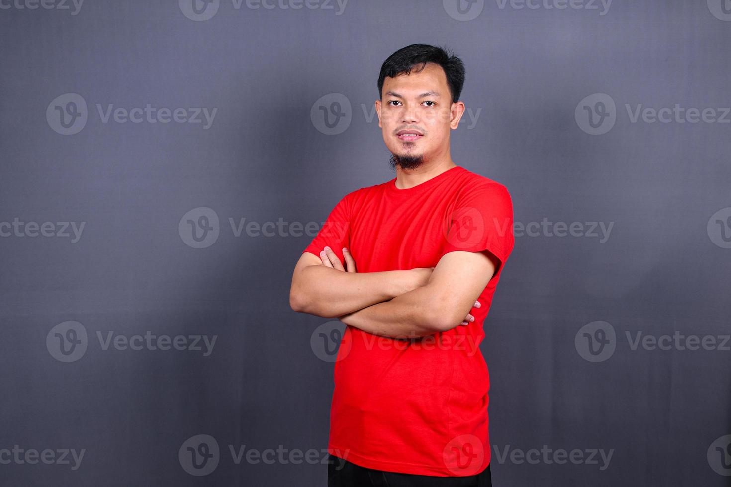 retrato de un atractivo hombre asiático con una camiseta roja de pie con los brazos cruzados aislado en un fondo gris foto