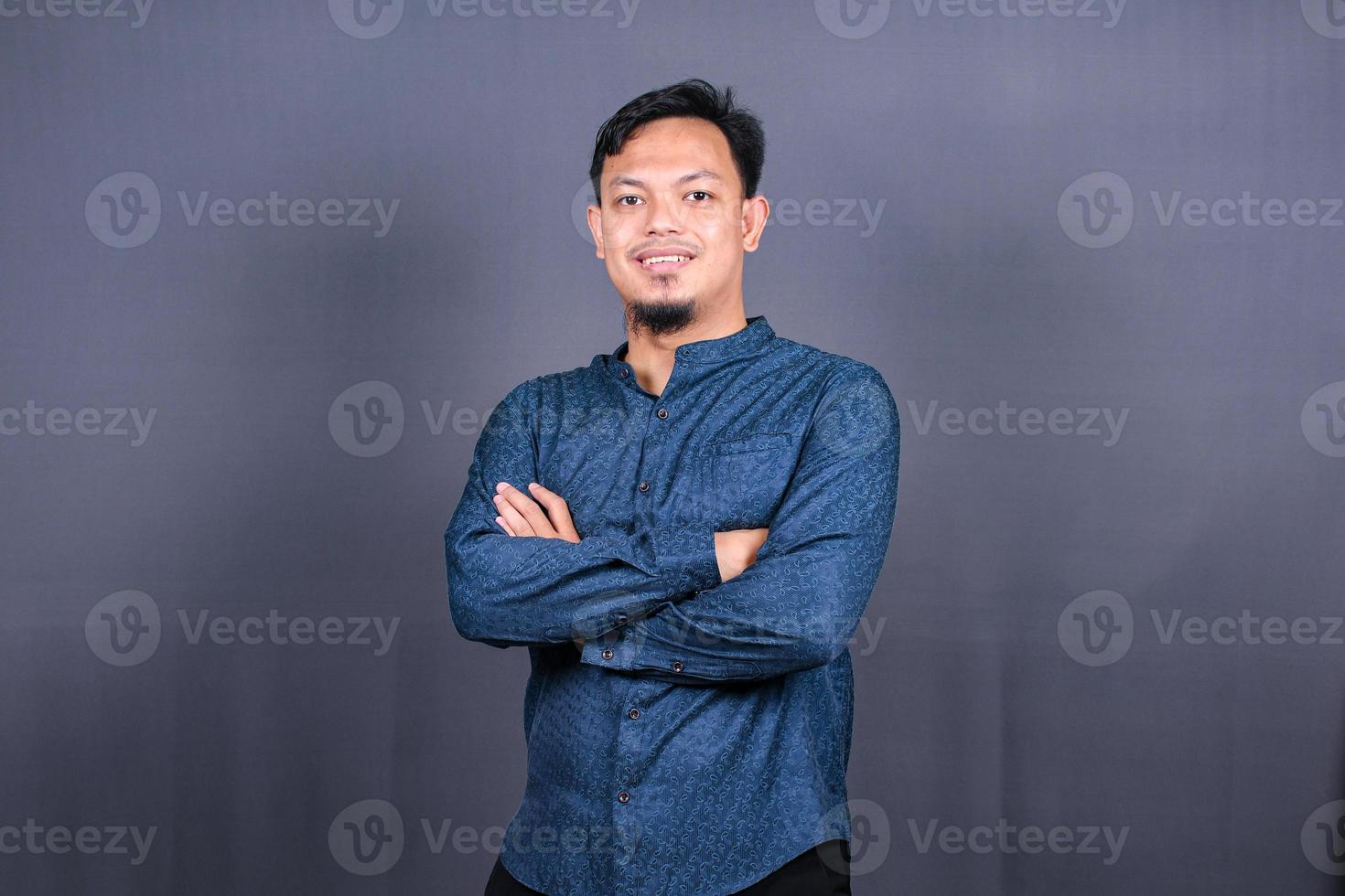 hombre asiático sonriente con camisa azul de pie con las manos dobladas aislado sobre fondo gris. concepto de trabajos de ingeniería foto