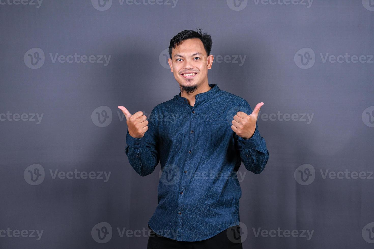 Young handsome man with blue shirt over gray background very happy and excited doing winner gesture with arms raised, smiling and screaming for success. Celebration photo
