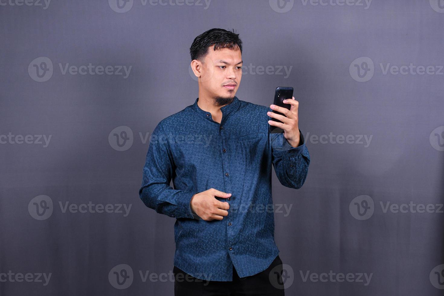 Portrait of unhappy Asian man in blue shirt holding mobile phone with sad expression on face. Isolated on gray background photo