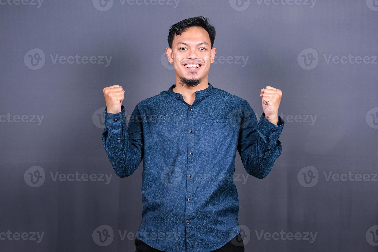 Young handsome man with blue shirt over gray background very happy and excited doing winner gesture with arms raised, smiling and screaming for success. Celebration photo