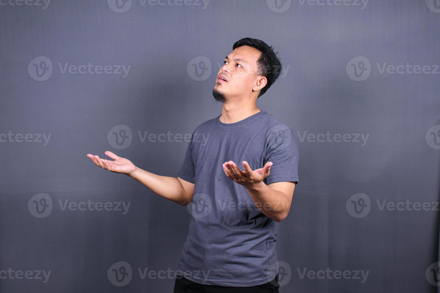 retrato de primer plano de un joven asiático de aspecto divertido y despistado, con los brazos abiertos preguntando qué hago ahora o gesticulando que no sé, aislado en un fondo gris. foto