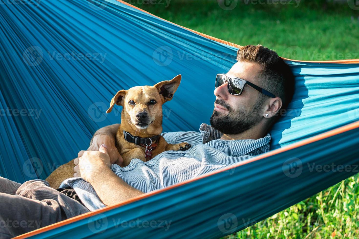 Smiling attractive European man in sunglasses is resting in hammock with his cute little dog. photo