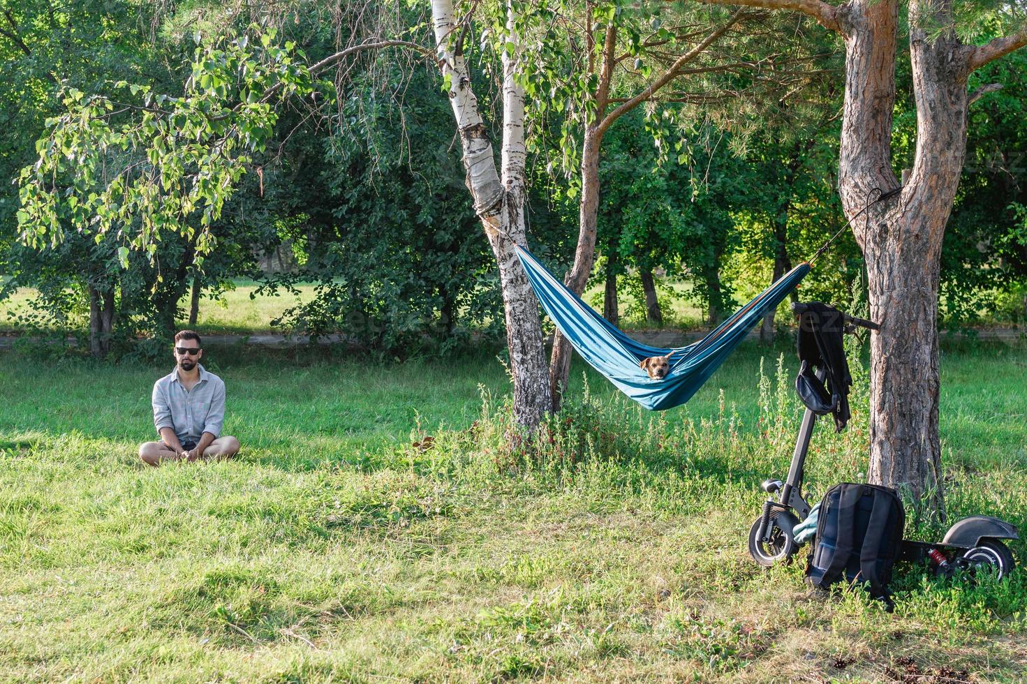 Handsome European man sitting on a grass in summer park near to hammock with dog, with electric scooter. photo
