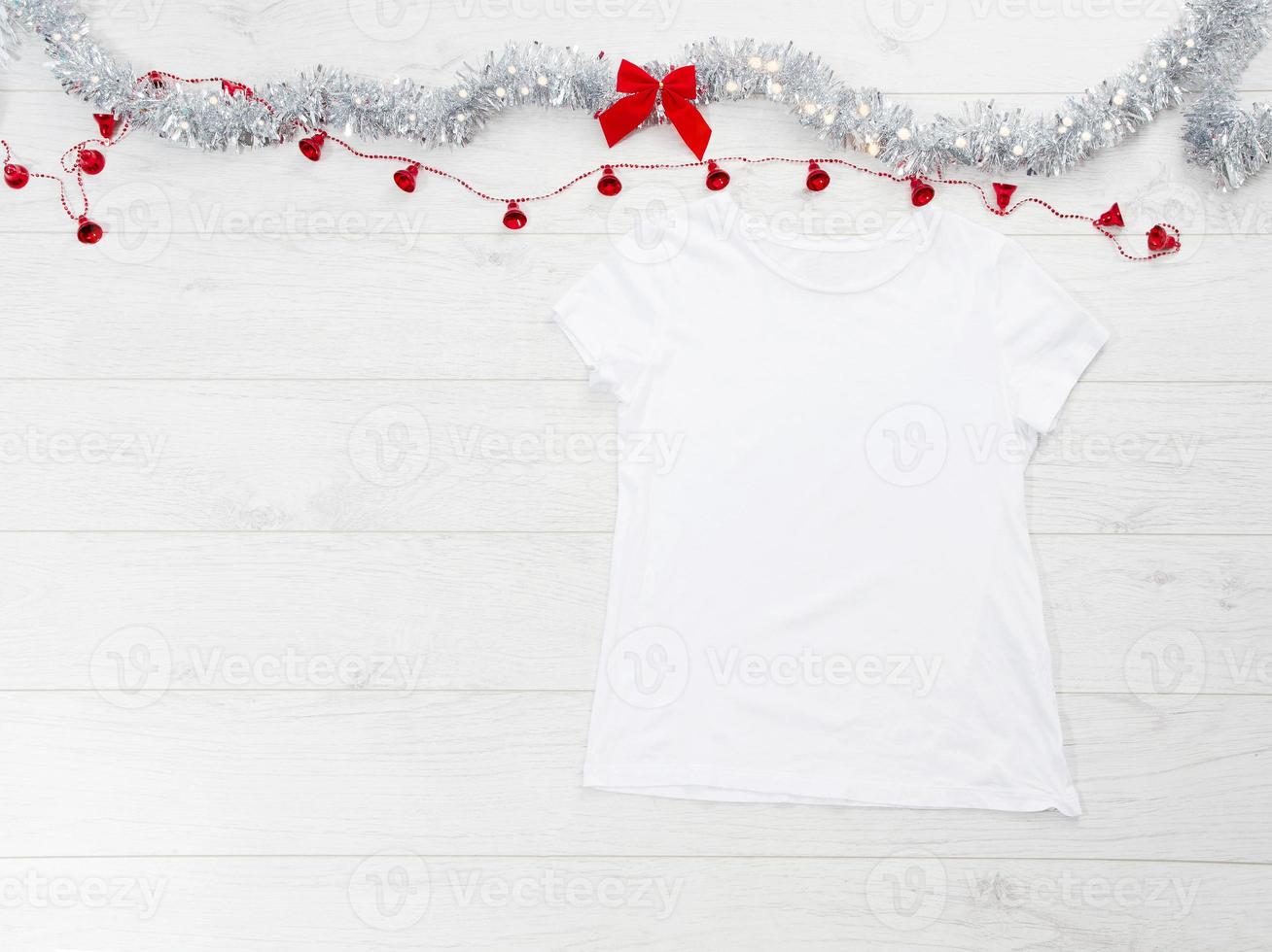 Close up white blank template t shirt with copy space and Christmas Holiday concept. Top view mockup t-shirt and red bow on white wooden background. Happy New Year decorations accessories. Xmas outfit photo