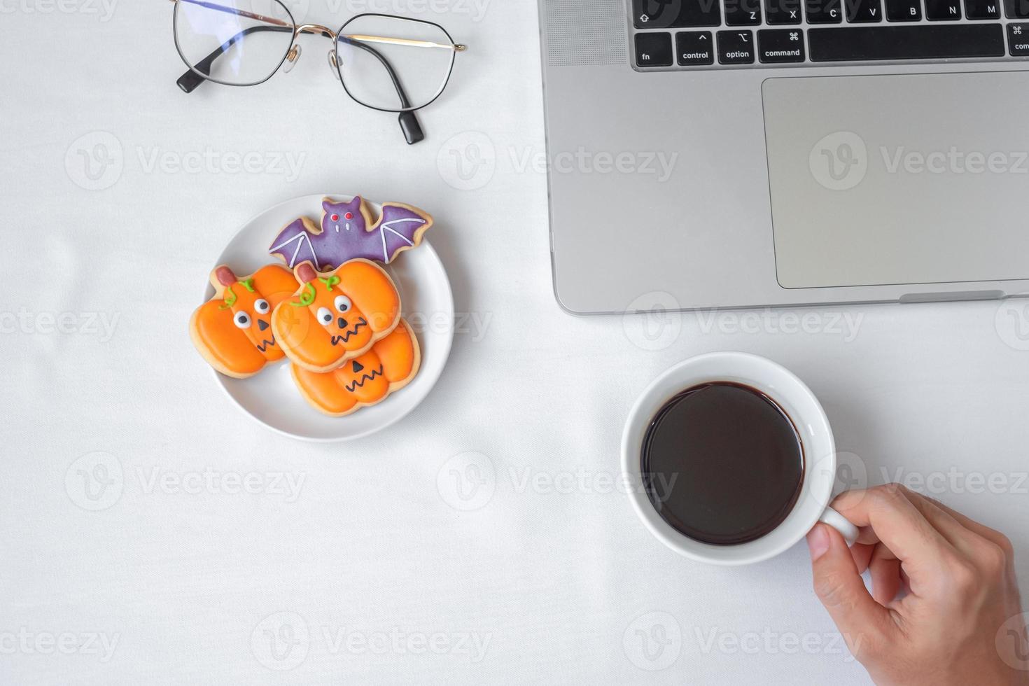 Hand holding coffee cup and eating Halloween Cookies during using computer laptop. Happy Halloween, online shopping, Hello October, fall autumn, Festive, party and holiday concept photo