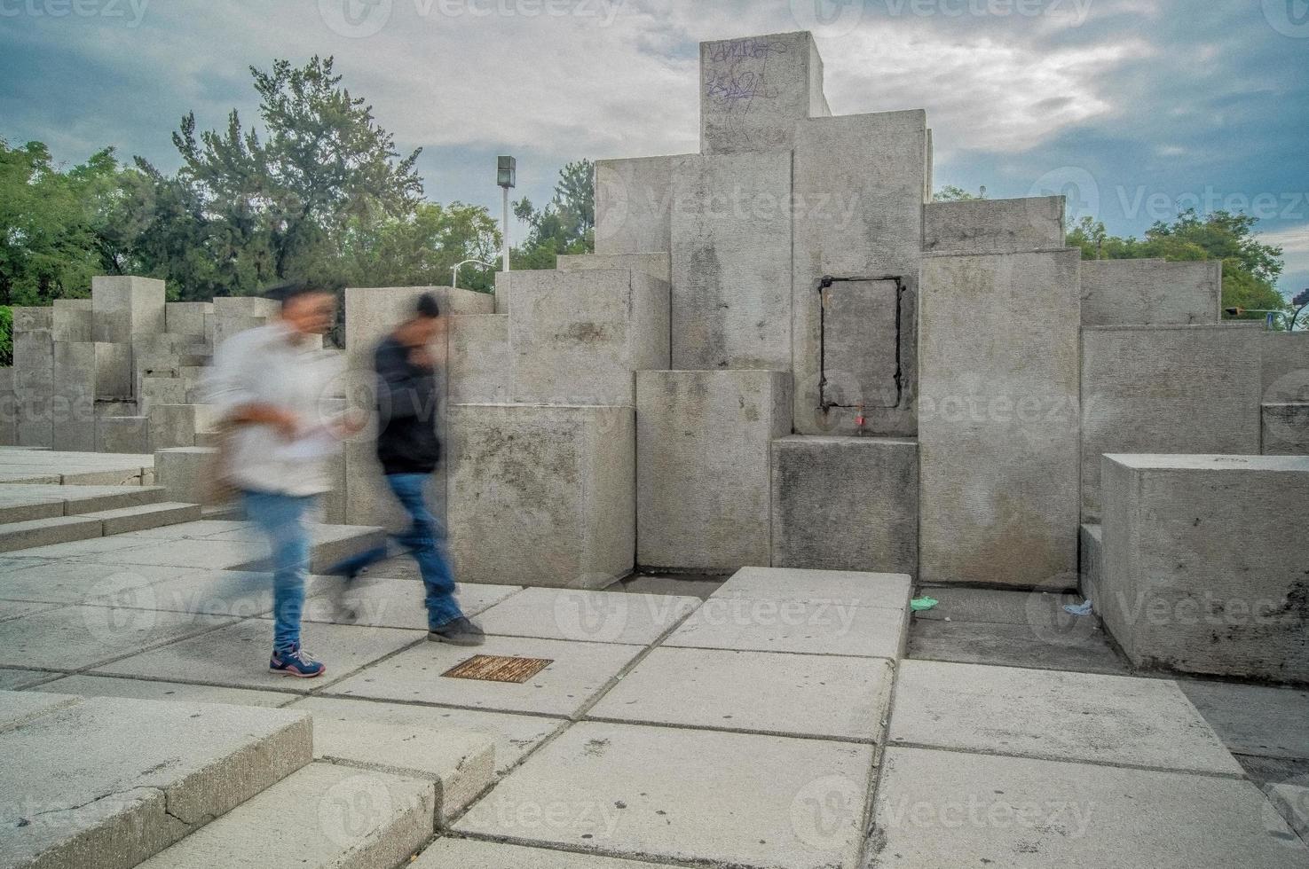 two people walking, long exposure blur, in the background stone sculpture different levels photo