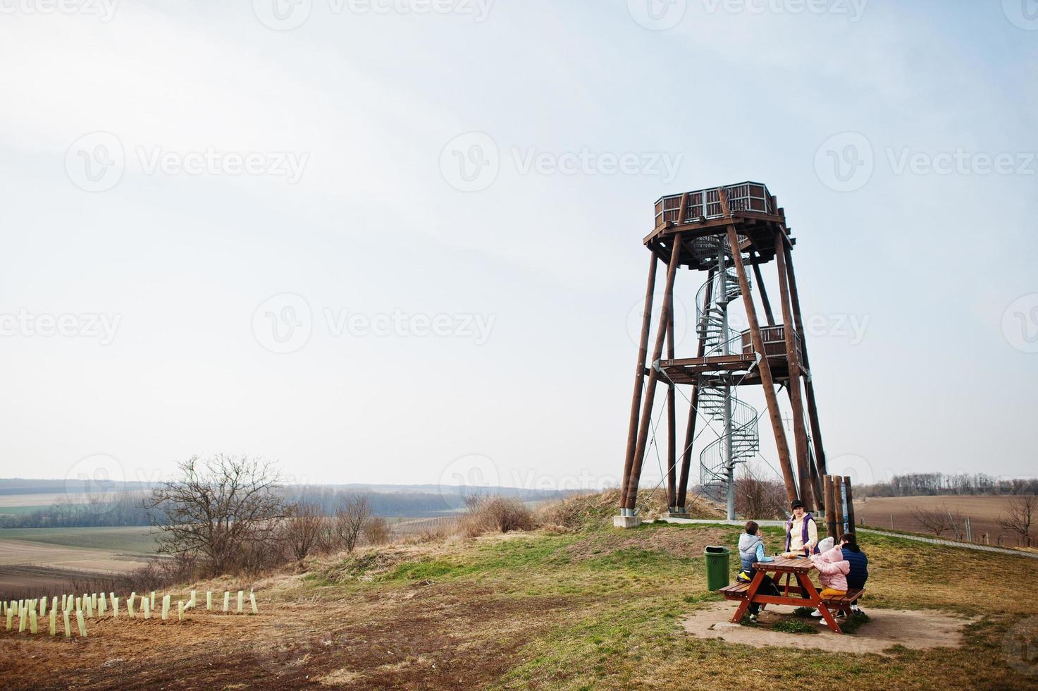 Mother with children sit at wooden table against observation tower. photo