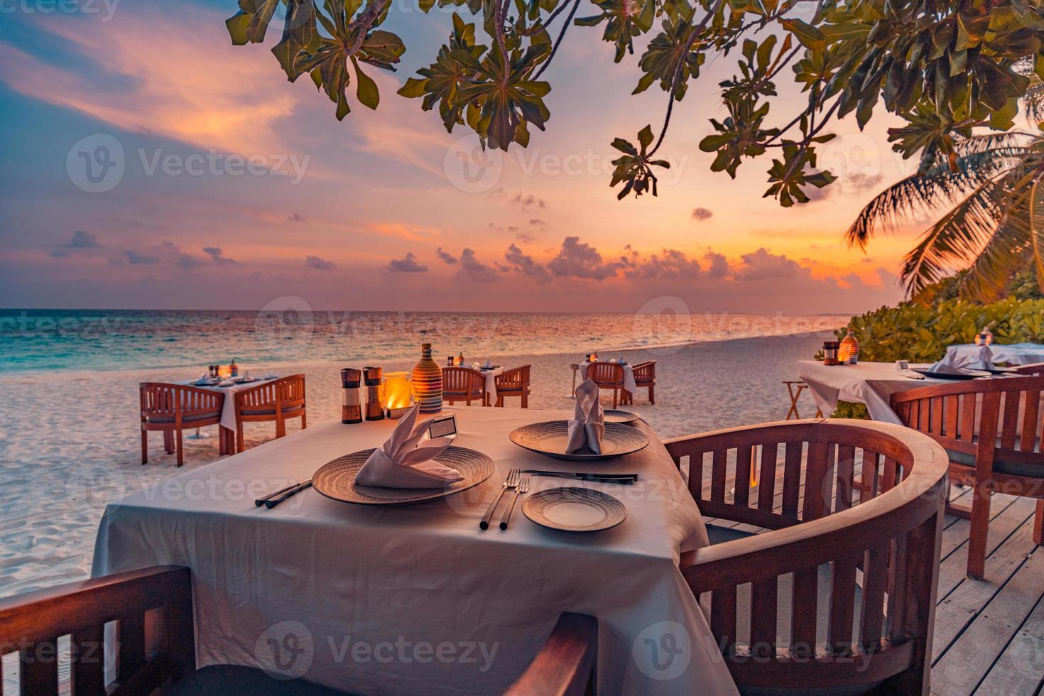 Outdoor restaurant at the beach. Table setting at tropical beach restaurant. Led light candles and wooden tables, chairs under beautiful sunset sky, sea view. Luxury hotel or resort restaurant photo