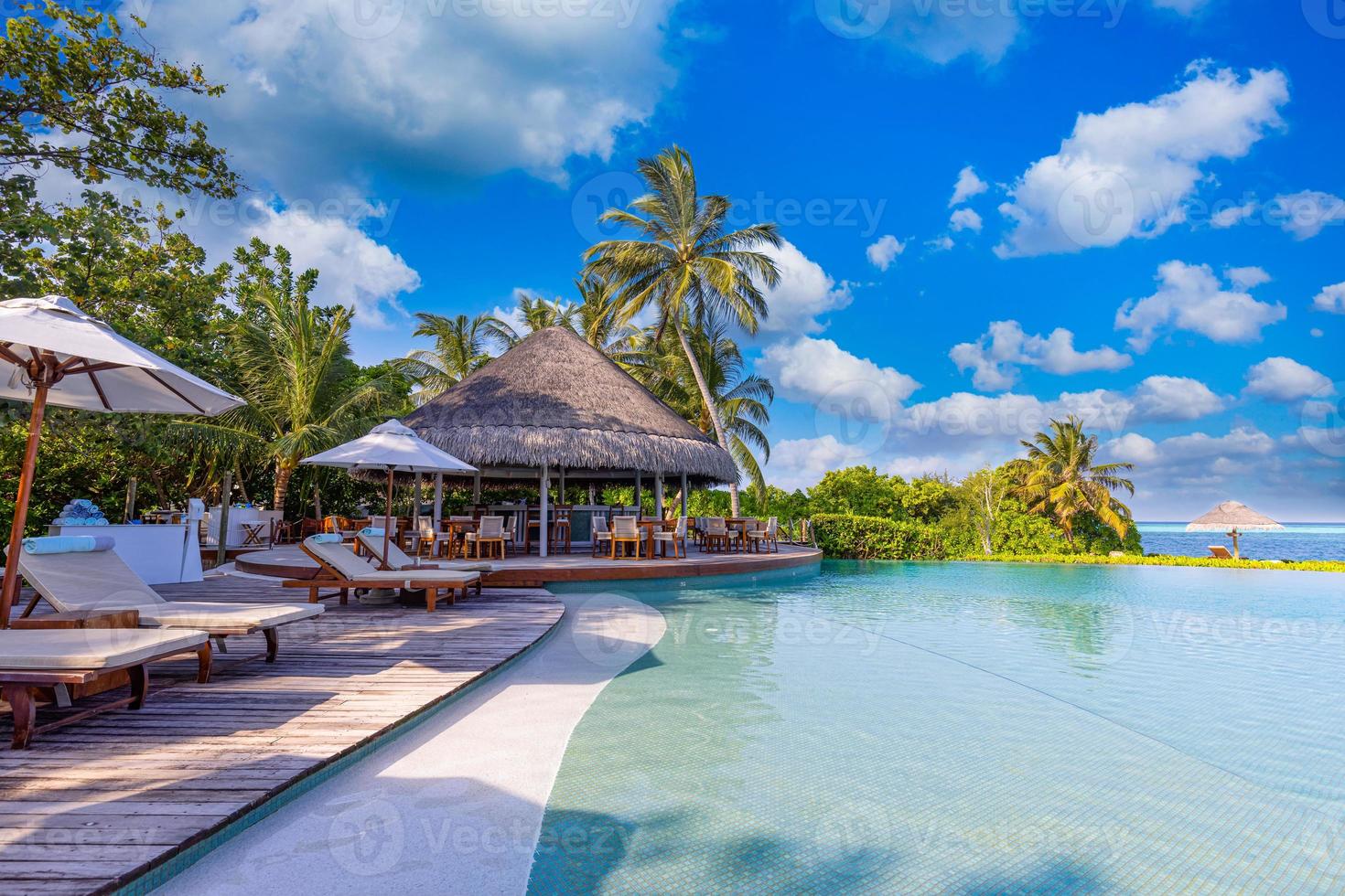 Recreational tourism landscape. Luxurious beach resort with spa swimming pool and beach chairs or leisure loungers under umbrellas with palm trees sky. Summer travel vacation background. Wellbeing photo