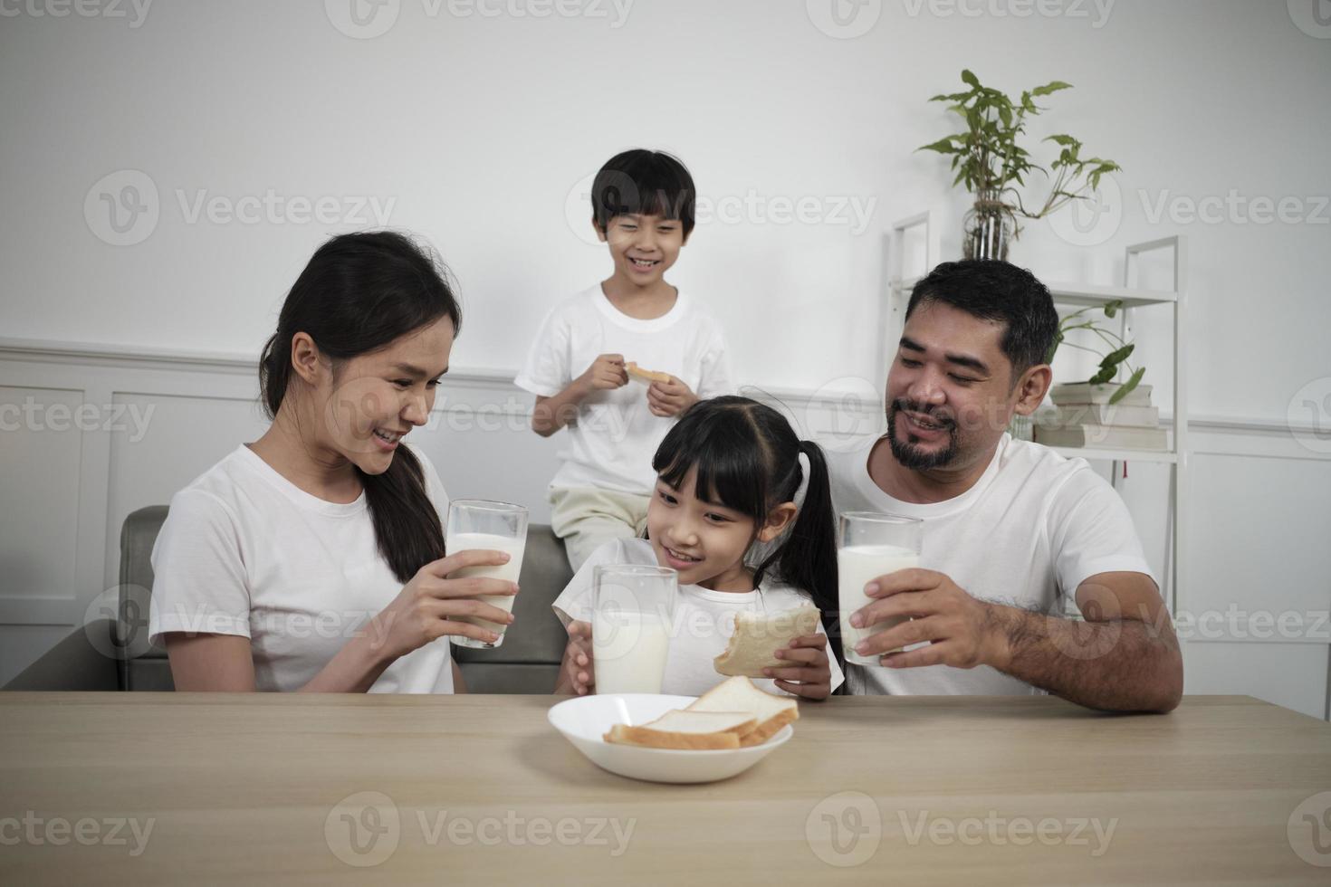 A Healthy Asian Thai family, little children, and young parents drink fresh white milk in glass and bread joy together at a dining table in morning, wellness nutrition home breakfast meal lifestyle. photo