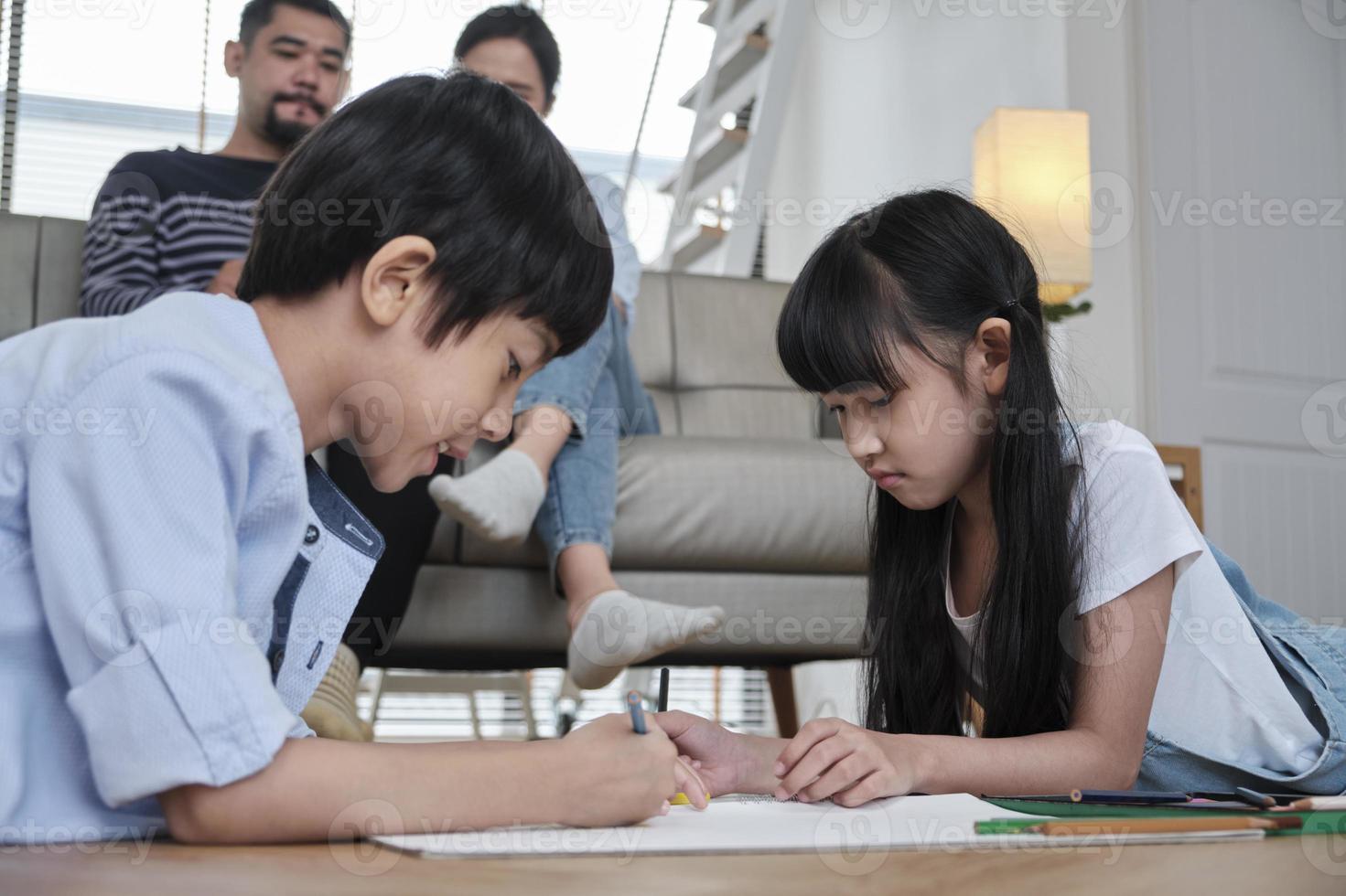 Asian Thai siblings are lying on living room floor, drawing homework with colored pencils together, parents leisurely relax on a sofa, lovely happy weekend activity, and domestic wellbeing lifestyle. photo