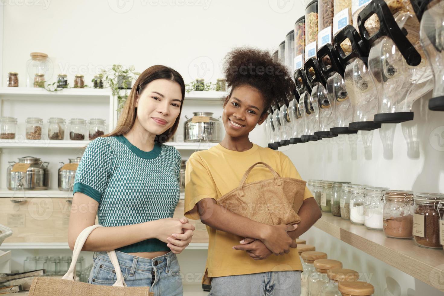 Two female customers look at camera and smile, shopping for organic products in refill store with reusable bags, zero-waste grocery, and plastic-free, environment-friendly, sustainable lifestyles. photo
