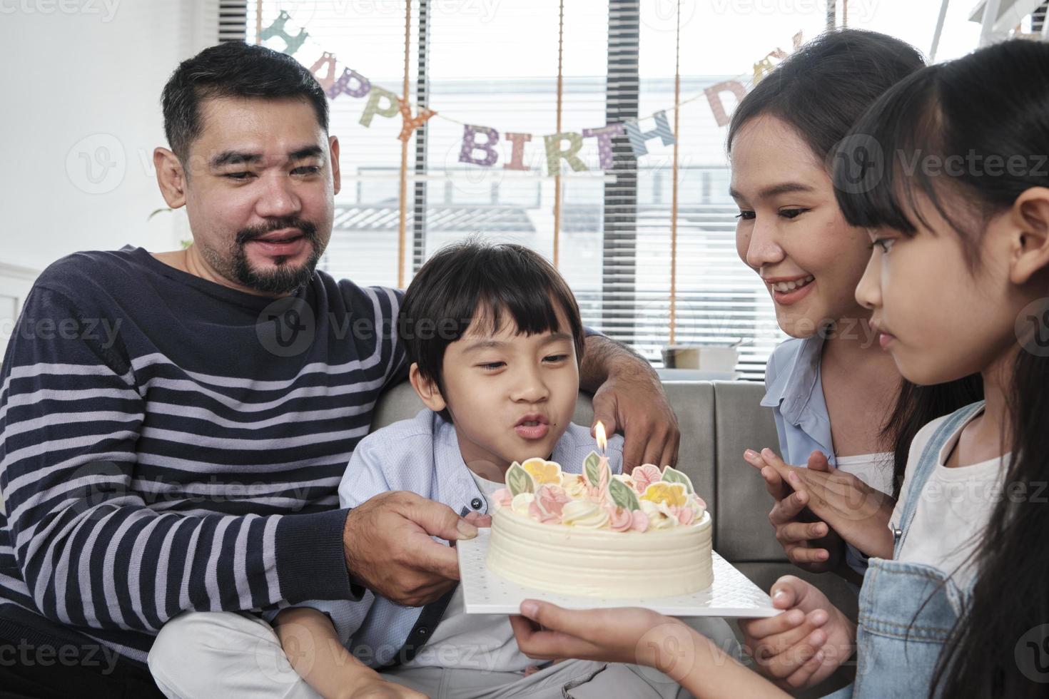 Happy Asian Thai family, young son is surprised with birthday cake, blows out candle, and celebrates joy party with parents and sister in living room together, wellbeing domestic home special event. photo