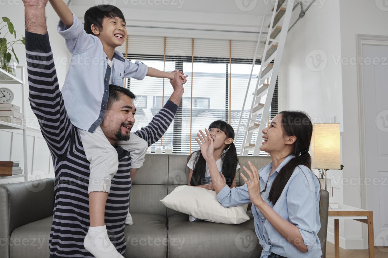 Asian Thai family together, dad plays and teases with children and mum by carrying and holding son on shoulders in home living room, happy leisure times, lovely weekend, wellbeing domestic lifestyle. photo