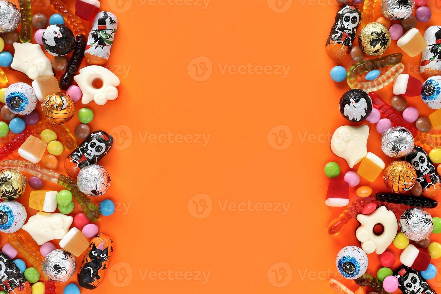 Halloweem orange background with copy space in middle, assorted candies on both sides - traditional eyeballs chocolates, jelly worms, ghosts. Happy Halloween holiday sale and trick-or-treat concept. photo