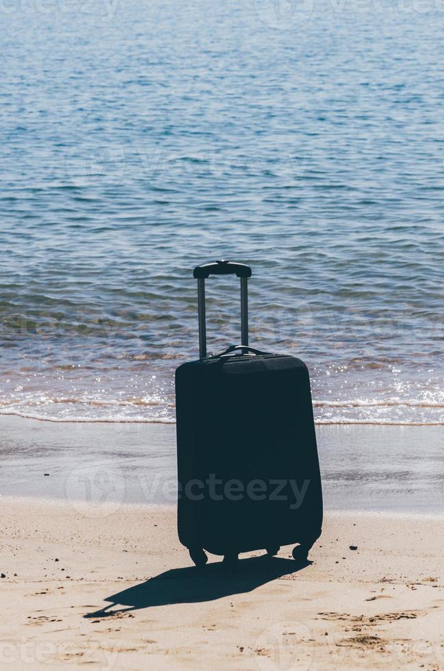 Black travel suitcase on sandy beach with turquoise sea background, summer holidays concept photo