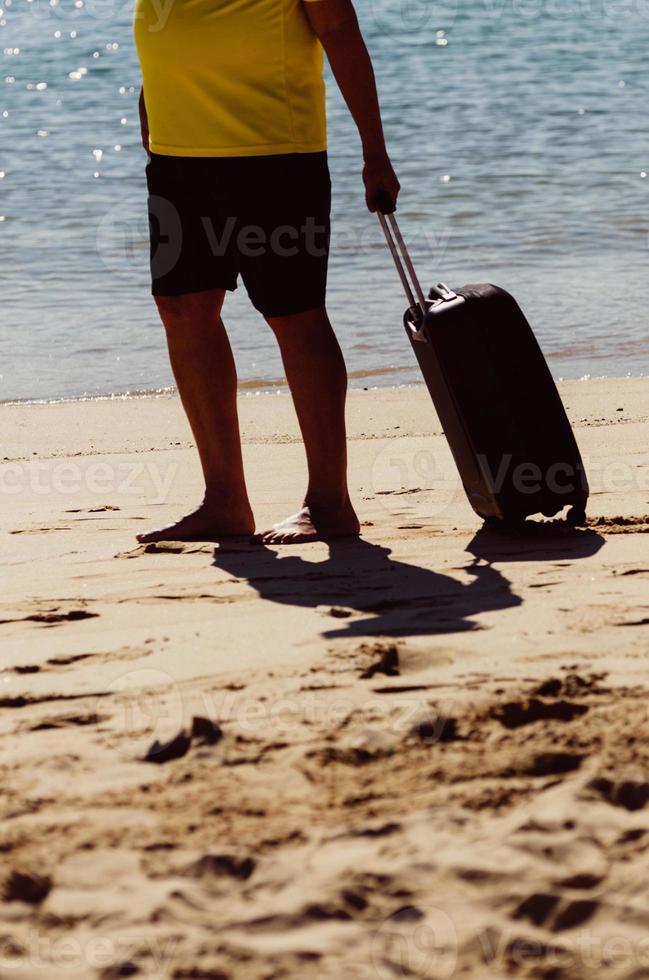 Barefood man wearing shorts and tshirt carrying a black travel suitcase on sandy beach with turquoise sea background, summer holidays concept photo