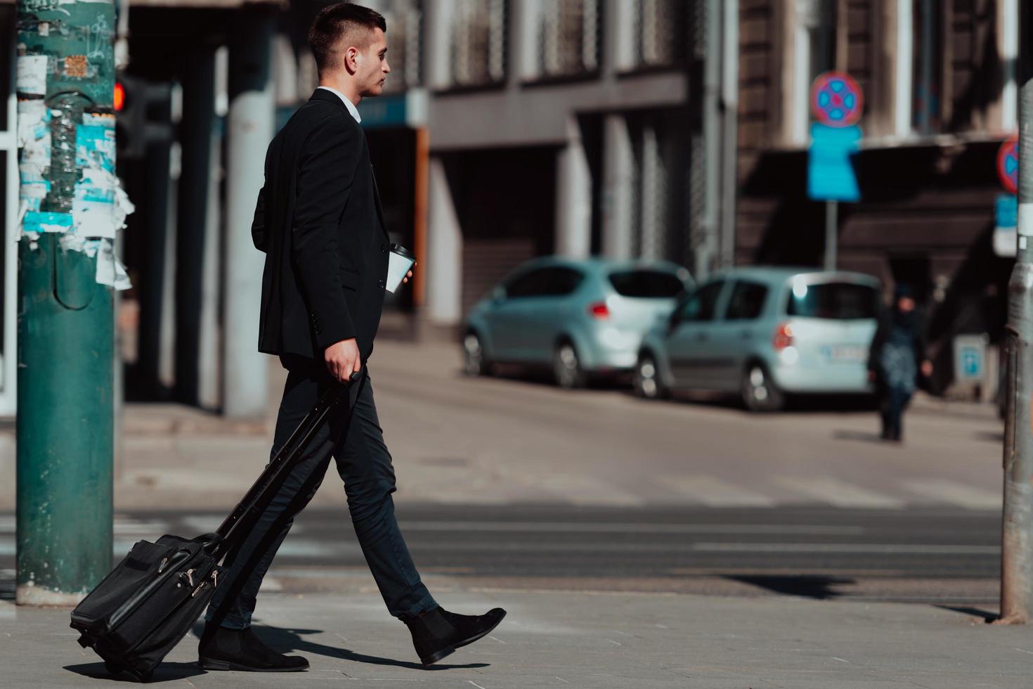 Going to airport terminal. Confident businessman traveler walking on city streets and pulling his suitcase drinking coffee and speaking on smartphone photo