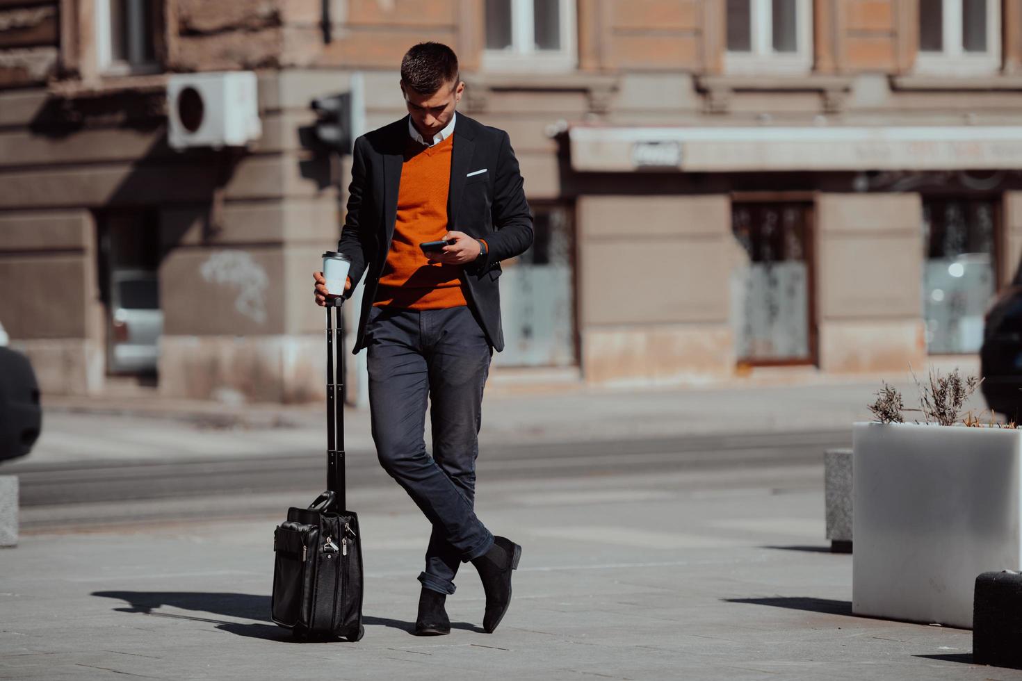 Going to airport terminal. Confident businessman traveler walking on city streets and pulling his suitcase drinking coffee and speaking on smartphone photo
