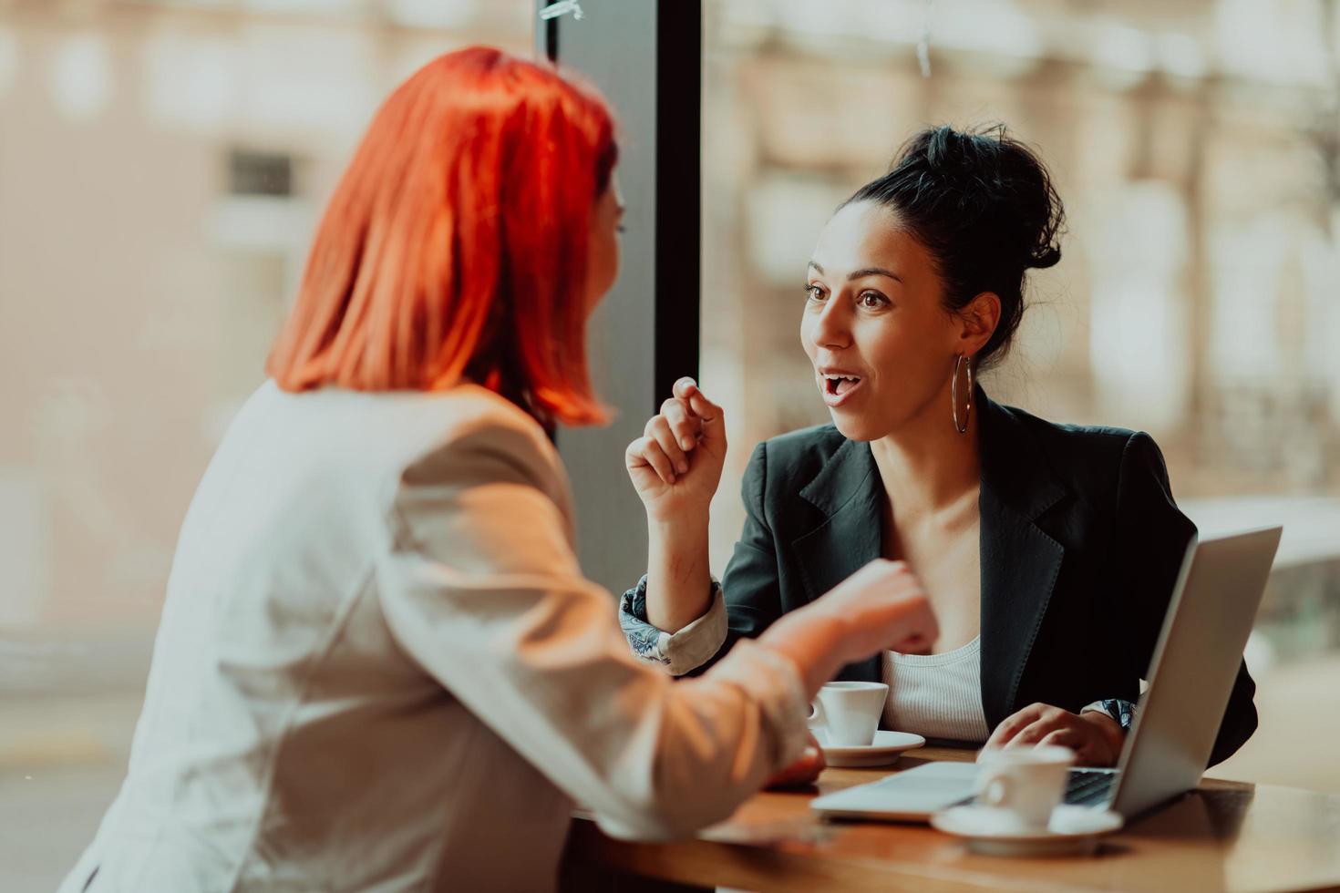 Two young business women sitting at table in cafe. Girl shows colleague information on laptop screen. Girl using smartphone, blogging. Teamwork, business meeting. photo