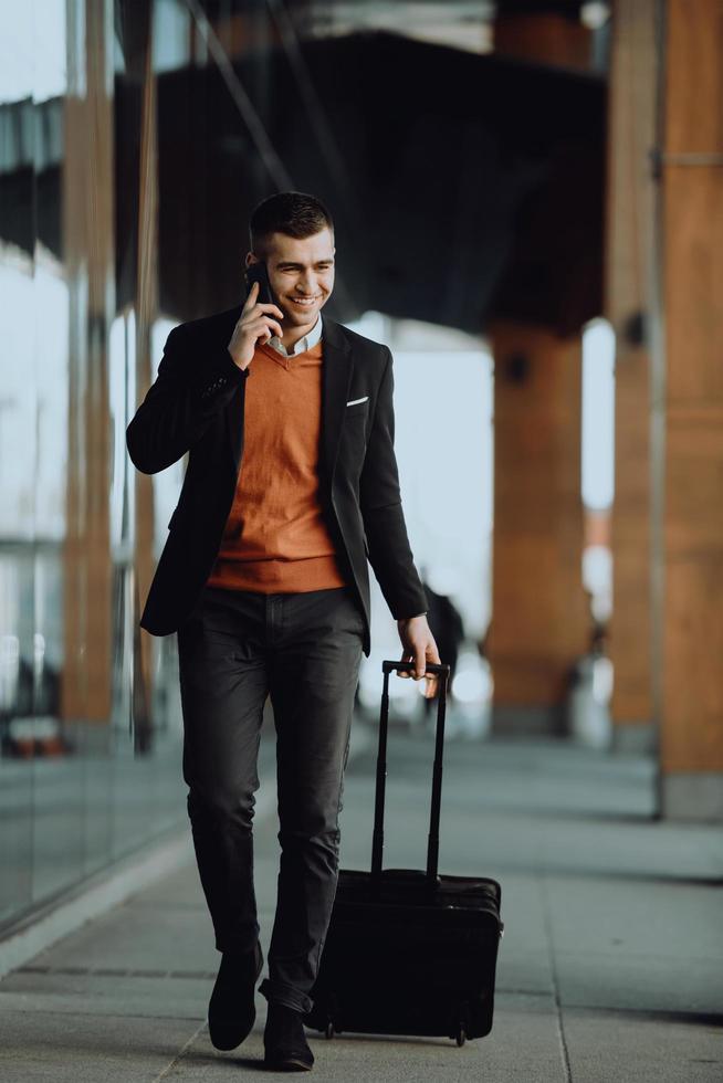 ir a la terminal del aeropuerto. viajero de negocios confiado caminando por las calles de la ciudad y tirando de su maleta bebiendo café y hablando en el teléfono inteligente foto