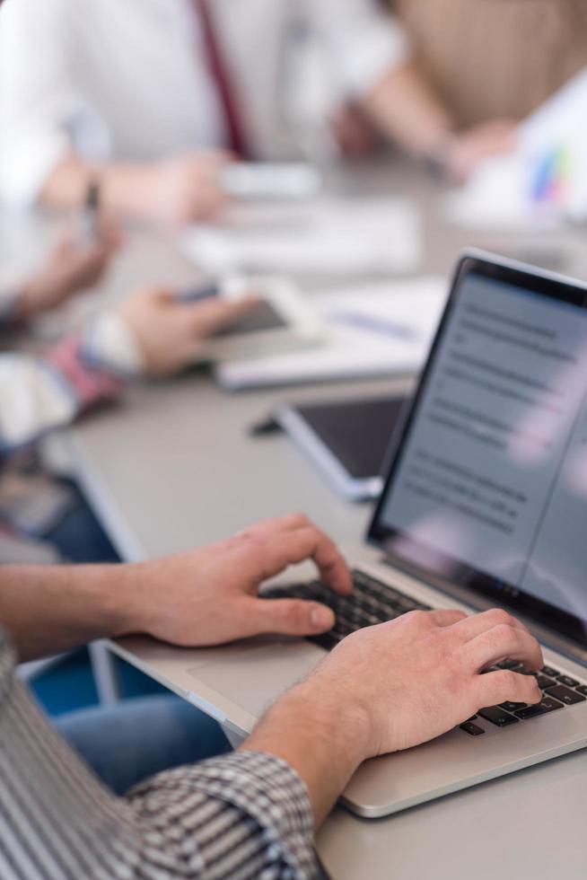close up of business man hands typing on laptop with team on meeting in background photo
